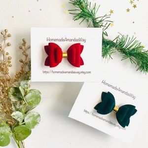May include: Two velvet hair bows, one red and one green, with gold centers. The bows are displayed on white cards with the text "HomemadeAmandasWay" and a website address.