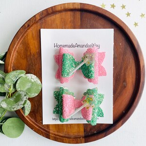 May include: Two glitter hair bows with a pink and green color scheme. The bows are decorated with a sparkly wand charm. The bows are displayed on a wooden tray with the text "HomemadeAmandasWay" and a website address.