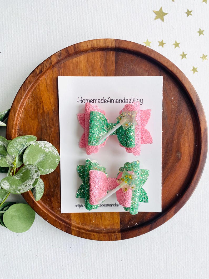 May include: Two glitter hair bows with a pink and green color scheme. The bows are decorated with a sparkly wand charm. The bows are displayed on a wooden tray with the text "HomemadeAmandasWay" and a website address.