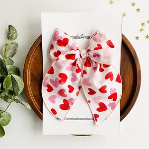 May include: A pink and white fabric bow with a red heart print. The bow is tied with a white ribbon and has a pink edge.
