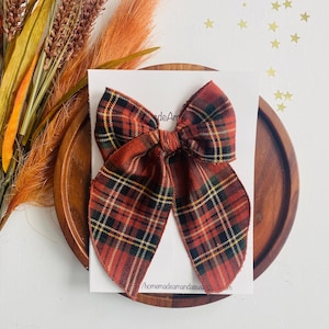 May include: A red, green, and yellow plaid bow with a frayed edge. The bow is tied in a knot and is sitting on a white card with the text "homemadeamandasugar" on it.