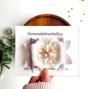 May include: A handmade hair bow with a large, pearlescent flower centre and pearl accents. The bow features a white lace design with iridescent glitter and lavender accents. The card reads "HomemadeAmandasWay".