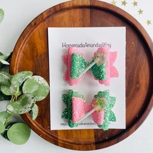 May include: Two green and pink glitter hair bows with a white lollipop stick in the center. The bows are on a white card with the text "HomemadeAmandasWay" and a website address.