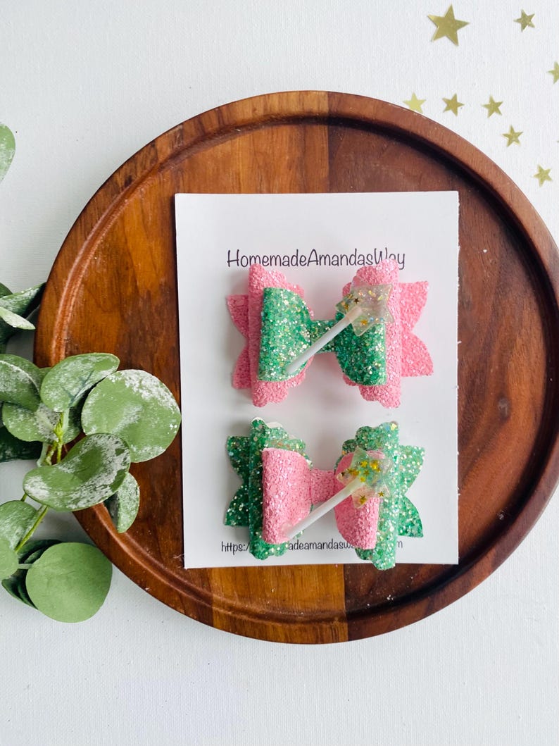 May include: Two green and pink glitter hair bows with a white lollipop stick in the center. The bows are on a white card with the text "HomemadeAmandasWay" and a website address.