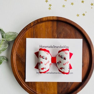 Puede incluir: Lazo rojo y blanco con estampado de Gato en el Sombrero. El lazo está sobre una superficie de madera con una tarjeta blanca que dice "HomemadeAmandasWay" y una dirección de sitio web.