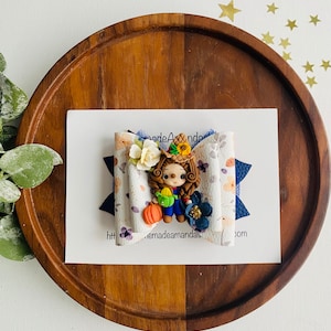 May include: A handmade fabric bow with a blue and white floral pattern. A small clay figurine of a girl wearing a blue dress and a straw hat is attached to the centre of the bow. The girl is holding a pumpkin and a watering can. The bow is on a wooden surface.