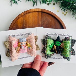 May include: Two glitter hair bows with a pink and gold bow featuring a miniature doll with a crown and a green and black bow featuring a miniature witch with a green hat and a broom. The bows are on white cards with the text "HomemadeAmandasWay" and a website address.