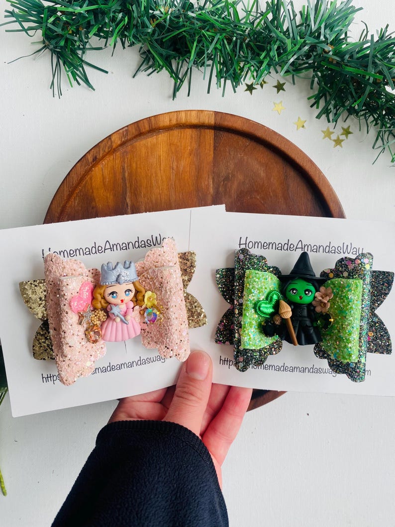 May include: Two glitter hair bows with a pink and gold bow featuring a miniature doll with a crown and a green and black bow featuring a miniature witch with a green hat and a broom. The bows are on white cards with the text "HomemadeAmandasWay" and a website address.