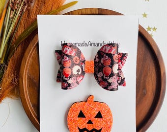 May include: A Halloween-themed hair bow with a black and orange pumpkin design. The bow is made of fabric with a glittery finish. The bow is attached to a clip. The bow is on a white background with a website address.