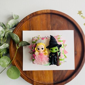 May include: A handmade polymer clay charm featuring a green witch with a black hat and a pink princess with a crown. The charm is on a green and pink patterned background.
