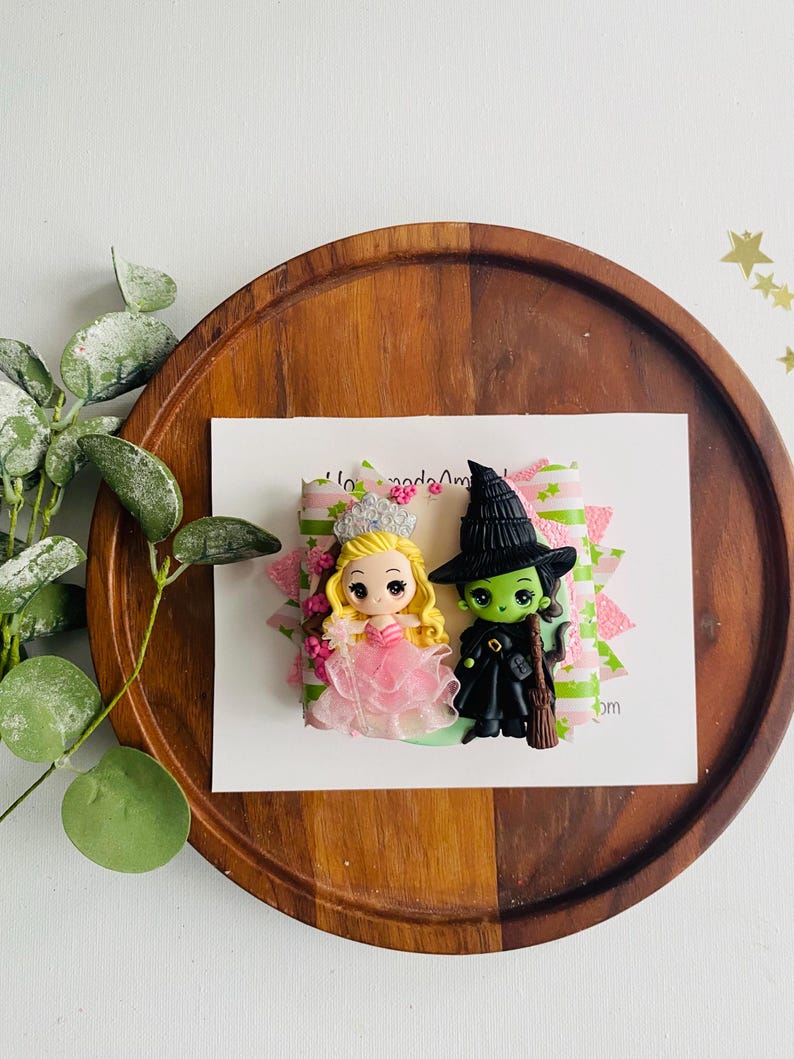 May include: A handmade polymer clay charm featuring a green witch with a black hat and a pink princess with a crown. The charm is on a green and pink patterned background.