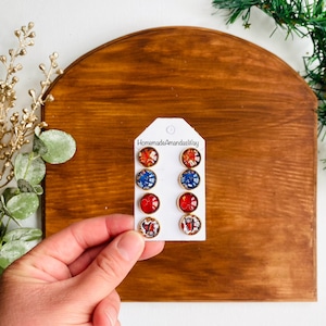 May include: A set of six pairs of earrings with a variety of designs featuring red, blue, and gold colors. The earrings are displayed on a white card with the text "HomemadeAmandasWay".