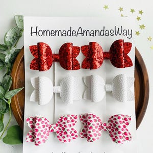 May include: Three sets of hair bows on a white card. The top row features red glitter bows, the middle row has white textured bows, and the bottom row displays bows with a pink strawberry pattern. The card reads "HomemadeAmandasWay".
