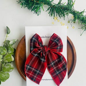 May include: A red and black plaid bow with a white background. The bow is tied with a knot in the center and has a slightly frayed edge. The bow is on a white background with the text "homemadeamandas" in black.