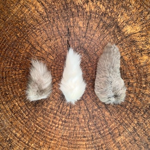 Puede incluir: Tres piezas de pelaje de conejo esponjoso en tonos grises y blancos, dispuestas sobre una superficie de madera marr&oacute;n texturizada. Las piezas de pelaje tienen forma de l&aacute;grima y un aspecto suave y afelpado.