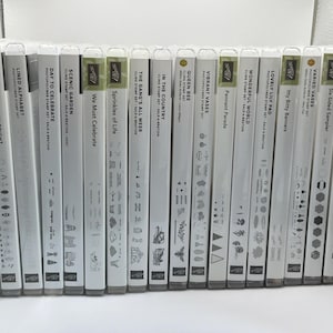 Puede incluir: Una colección de diecisiete estuches de plástico transparente para sellos dispuestos en fila. Cada estuche presenta un lomo blanco con texto negro que identifica el nombre del set, como Vintage Christmas, Queen Bee y Varied Vases, junto con pequeños iconos gráficos negros.