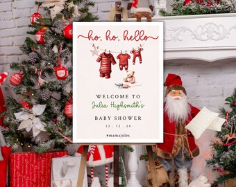 May include: A white sign with red and green text that says "ho, ho, hello" and "Welcome to Julia Highsmith's Baby Shower 12.12.24 #mamajules". There are illustrations of baby clothes hanging on a clothesline and a teddy bear sitting on the ground. The sign is propped up on a wooden easel in front of a Christmas tree and a Santa Claus figurine.