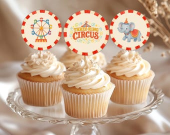 May include: Cupcakes with white frosting and decorative toppers featuring a Ferris wheel, an elephant, and the text "THREE-RING CIRCUS." The cupcakes are displayed on a glass cake stand, creating a festive and celebratory atmosphere.