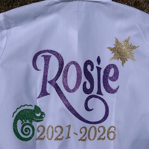 May include: White button-down shirt featuring a purple glitter "Rosie" design, a gold glitter sun, a green glitter chameleon, and the gold text "2021-2026". The shirt is hanging on a black hanger.