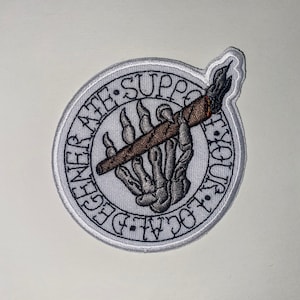 May include: Embroidered patch featuring a skeletal hand holding a lit cigar. The patch is circular with a white border and text that reads "LOCAL LEGEND ATE SUPPORT". The cigar has a brown body with orange and black details.