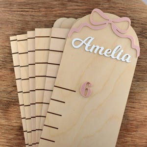 May include: A light-colored wooden growth chart featuring a rounded top with a pink bow cutout. The name Amelia is written in white script lettering, and the number 6 is displayed in pink. The chart includes horizontal black measurement lines.