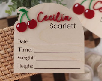 May include: A light-colored wooden arch-shaped birth announcement sign featuring a red acrylic cherry graphic and the names Cecilia Scarlett in red and black text. The sign includes printed lines for Date, Time, Weight, and Height details.