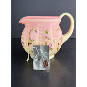 May include: A vintage pink and yellow Fenton pitcher with a floral design. The pitcher has a curved handle and a spout. A small tag with a black and white image is attached to the pitcher.