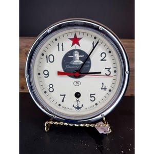 May include: A vintage round clock with a silver-colored bezel and a white face. The clock features black numerals, a red star, and a small anchor symbol. The clock is mounted on a decorative gold-colored stand.