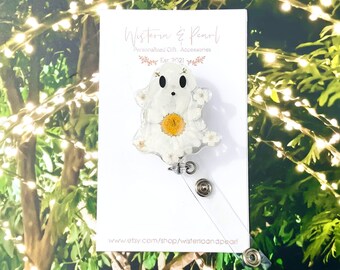 Puede incluir: Un portagafete blanco en forma de fantasma con un diseño de flores blancas y un centro de flor amarillo. El portagafete está en una tarjeta blanca con el texto "Wisteria & Pearl Personalized Gift Accessories Est 2021" y una dirección de sitio web.