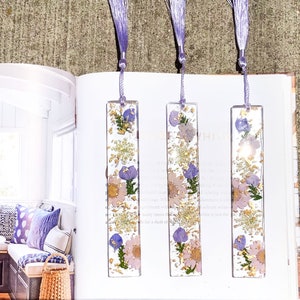 May include: Three purple and white floral bookmarks with gold leaf accents. The bookmarks are made of clear resin and have a tassel on the top.