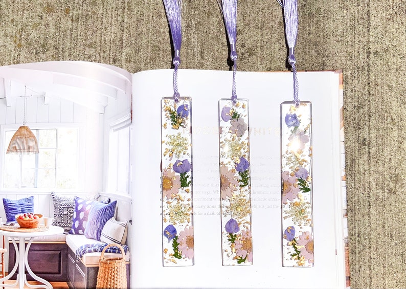 May include: Three purple and white floral bookmarks with gold leaf accents. The bookmarks are made of clear resin and have a tassel on the top.