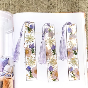 May include: Three purple and gold resin bookmarks with pressed flowers and a purple tassel.