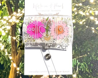 May include: A clear, rectangular badge reel with a floral design. The badge reel features pressed pink and orange flowers, with a heart-shaped arrangement of flowers in the center. The badge reel has a retractable cord and a metal clip.