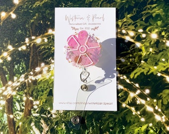 May include: Pink and white floral resin badge reel with a white retractable cord and a silver clip. The badge reel is on a white card with the text "Wisteria & Pearl Personalised Gift Accessories Est. 2021" and the website "www.etsy.com/shop/wisteriaandpearl".