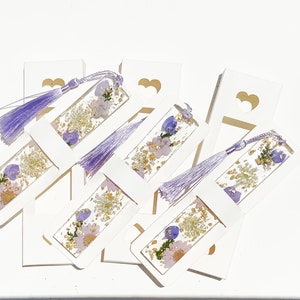May include: A set of six white bookmarks with purple tassels. Each bookmark features a pressed flower design with purple and gold accents.