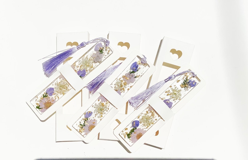 May include: A set of six white bookmarks with purple tassels. Each bookmark features a pressed flower design with purple and gold accents.