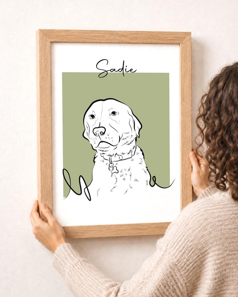 May include: A framed art print featuring a line drawing of a dog with the name "Sadie" above. The dog is white with black outlines, set against a muted green background. The frame is made of light-colored wood.