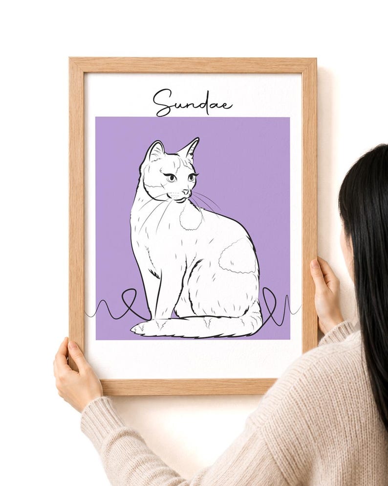 May include: A framed art print featuring a white cat illustration on a lavender background. The cat is drawn in a minimalist style with black outlines and a few spots. The word "Sundae" is written above the cat in cursive.