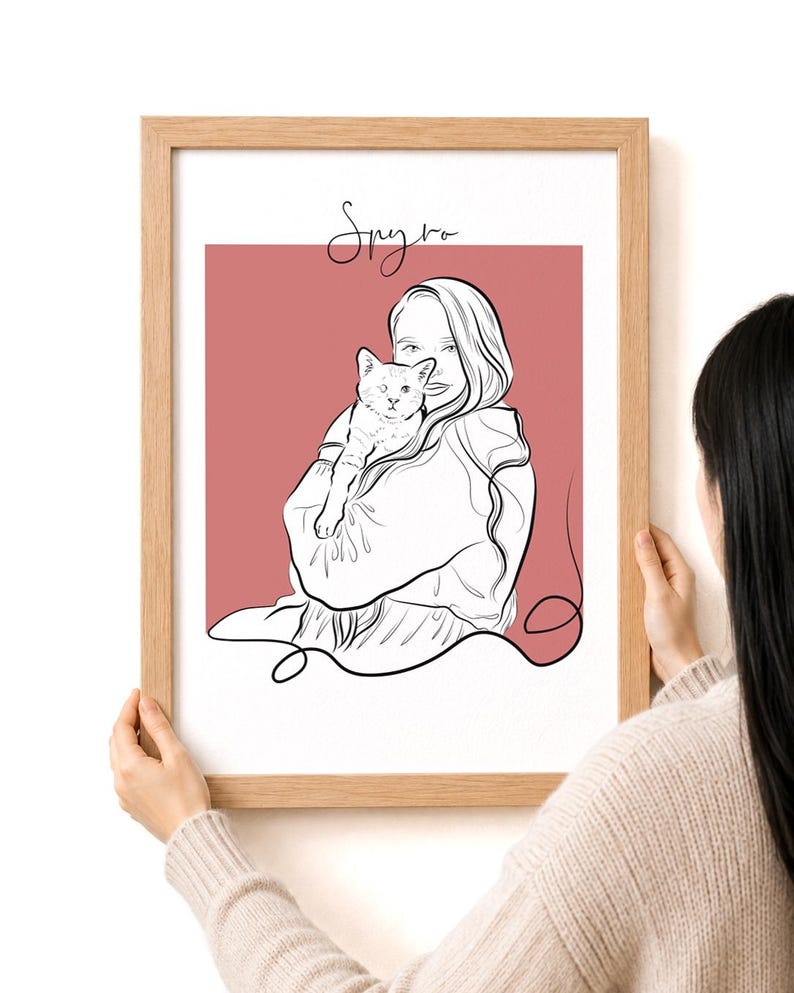 May include: A framed line art print featuring a person holding a cat. The artwork is in black and white, set against a dusty rose background. The frame is made of light-colored wood, and the print has the word "Smyrka" in cursive.