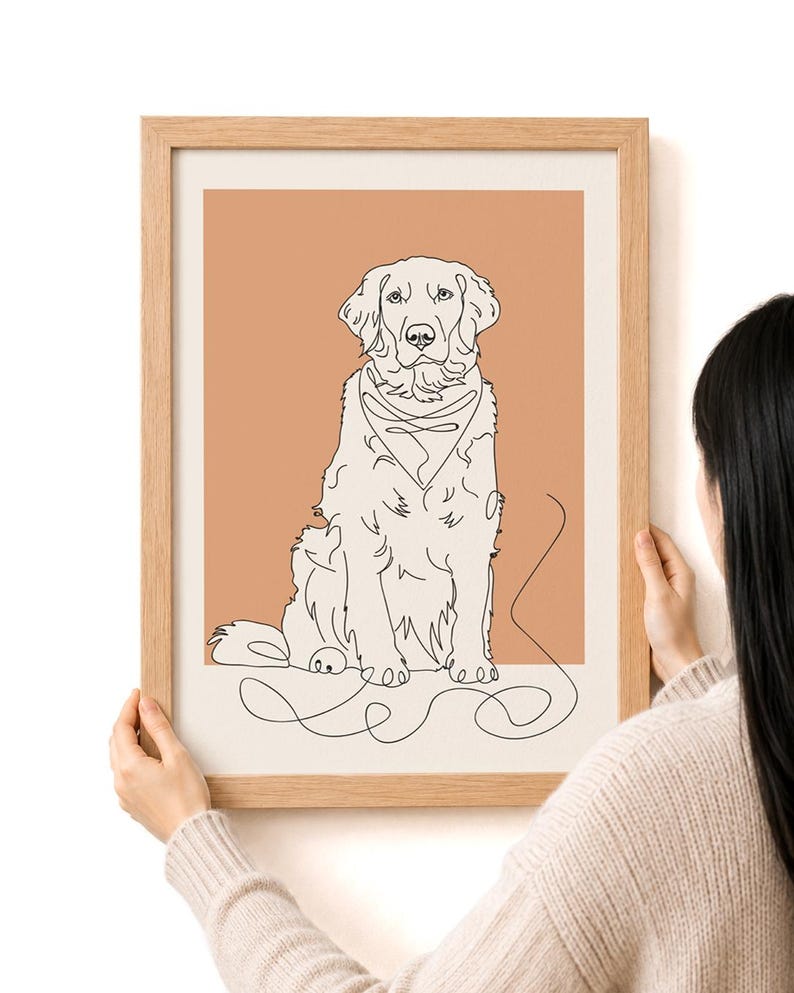 May include: A framed art print featuring a line drawing of a Golden Retriever dog. The dog is depicted in a sitting position with a bandana, set against a peach-colored background. The frame is made of light-colored wood.