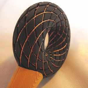 Può includere: Una scultura nera a forma di toro con un motivo geometrico di linee color rame. La scultura è montata su un manico di legno marrone chiaro. Il design presenta un motivo curvo e ripetitivo, creando un pezzo visivamente interessante.