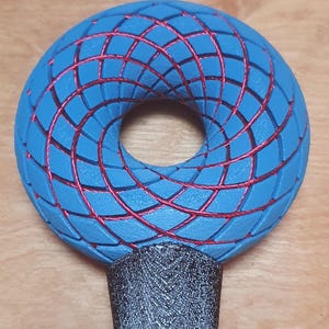 Puede incluir: Una escultura geométrica azul y roja con un diseño circular. La escultura tiene una superficie azul en forma de rejilla con líneas rojas que la atraviesan. La base es de color gris oscuro.