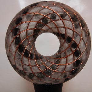 May include: A donut-shaped sculpture with a central hole. The sculpture is made of a translucent material with black and grey inclusions, and is overlaid with a copper wire grid. The base is black.