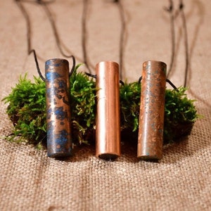May include: Three cylindrical metal pendants with different finishes. The pendants are hanging from black cords and are displayed on a bed of green moss. The pendants are a blue-green, copper, and a rusty brown.