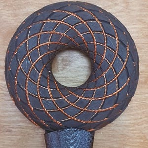 Könnte beinhalten: Dunkelgraues, kreisförmiges Objekt mit einem zentralen Loch, das ein gewebtes Kupferdrahtdesign aufweist. Das Objekt ist auf einem strukturierten, dunkelgrauen Griff montiert. Das Design ist symmetrisch und geometrisch.