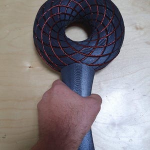 May include: A dark gray, torus-shaped object with a handle. The torus is constructed with a grid of red wires. The handle is cylindrical and gray. The object is held by a hand.