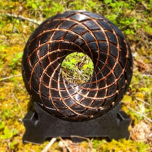 May include: A black spherical object with a hollow centre and a textured, grid-like surface pattern featuring copper-coloured lines. The object is mounted on a black, angular stand that supports its circular form in an upright position.