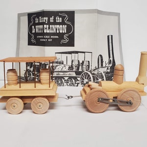 May include: A wooden model kit of the De Witt Clinton steam locomotive, a historic American train. The kit includes a wooden train engine and a wooden boxcar with barrels. The kit comes with instructions and is ready to be assembled.