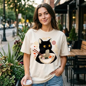 Peut inclure: Un t-shirt à manches courtes de couleur crème arborant un motif de chat noir mangeant des nouilles dans un bol avec des baguettes. Le design comprend des nuages stylisés et un emblème rouge circulaire. Le t-shirt a un col rond.