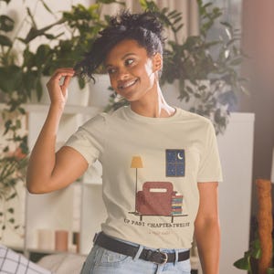 Peut inclure: Un t-shirt à manches courtes de couleur crème avec un motif représentant une pièce avec un fauteuil, une lampe, une fenêtre avec un croissant de lune et une pile de livres. Le texte indique UP PAST CHAPTER TWELVE et a night reader.